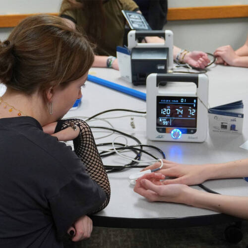 A student's blood pressure and pulse oximetry readings display on a medical monitor as a pulse oximeter is clipped to a classmate's finger, part of a hands-on healthcare or medical science activity around a shared table.